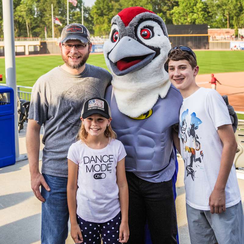 Family posing with a baseball team mascot at a stadium, smiling during a game-day event