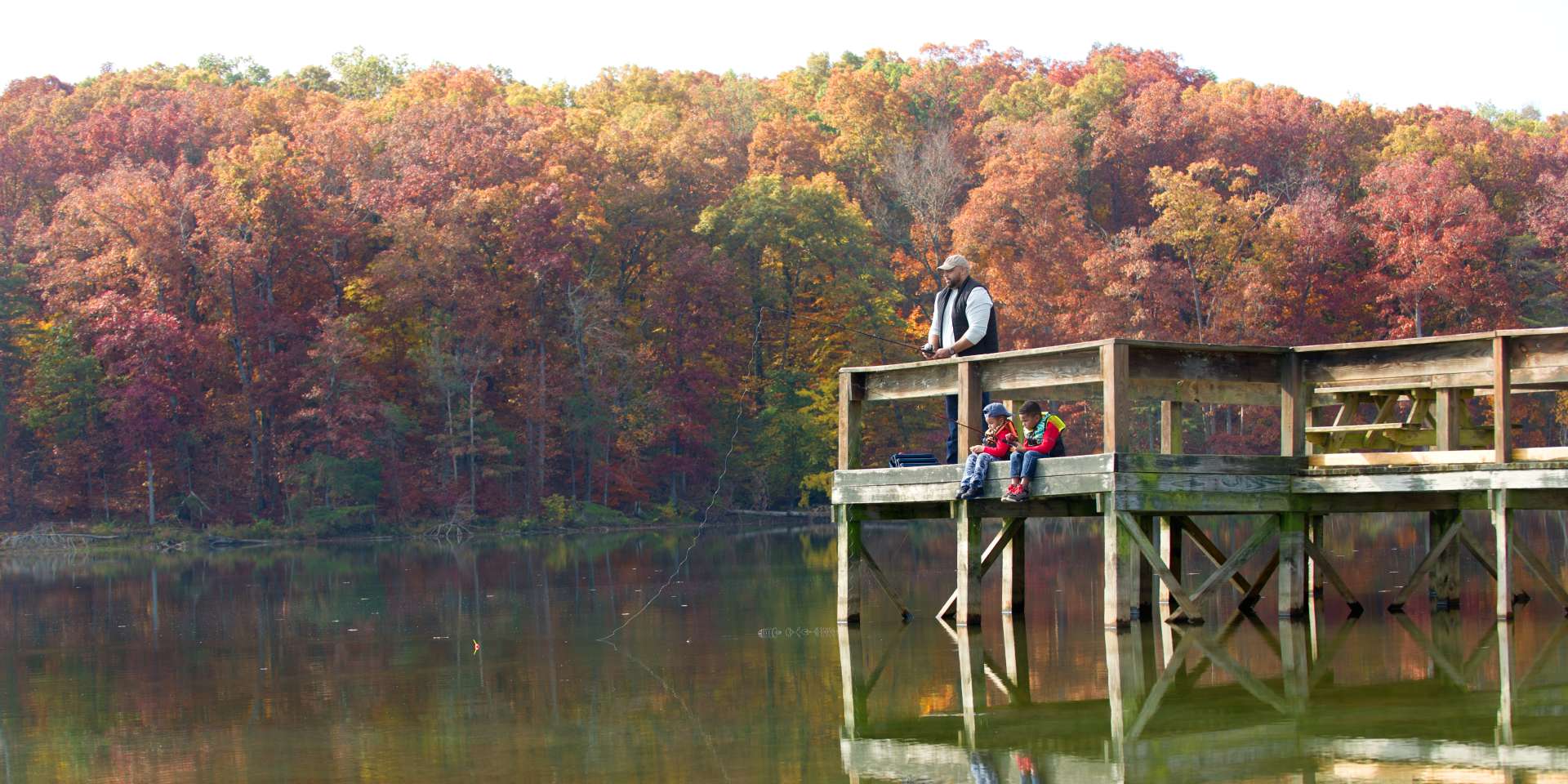 Two boys with their dad fishing on a dock surrounded by fall foliage at Deam Lake State Recreation Area