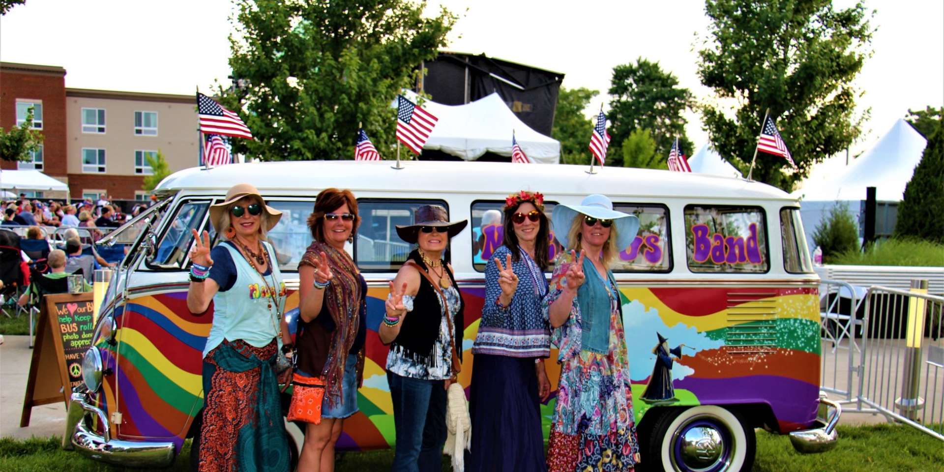 five women in hippie clothing giving peace sign stand in front of a brightly painted VW bus
