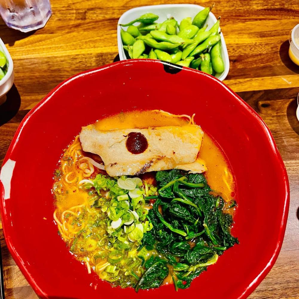 A bowl of spicy chicken ramen in a red bowl sits on a wood table next to a smaller bowl of edamame.