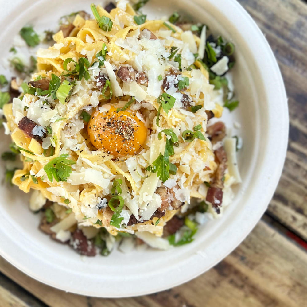Heavy-duty paper plate on a wooden table, full of carbonara pasta.