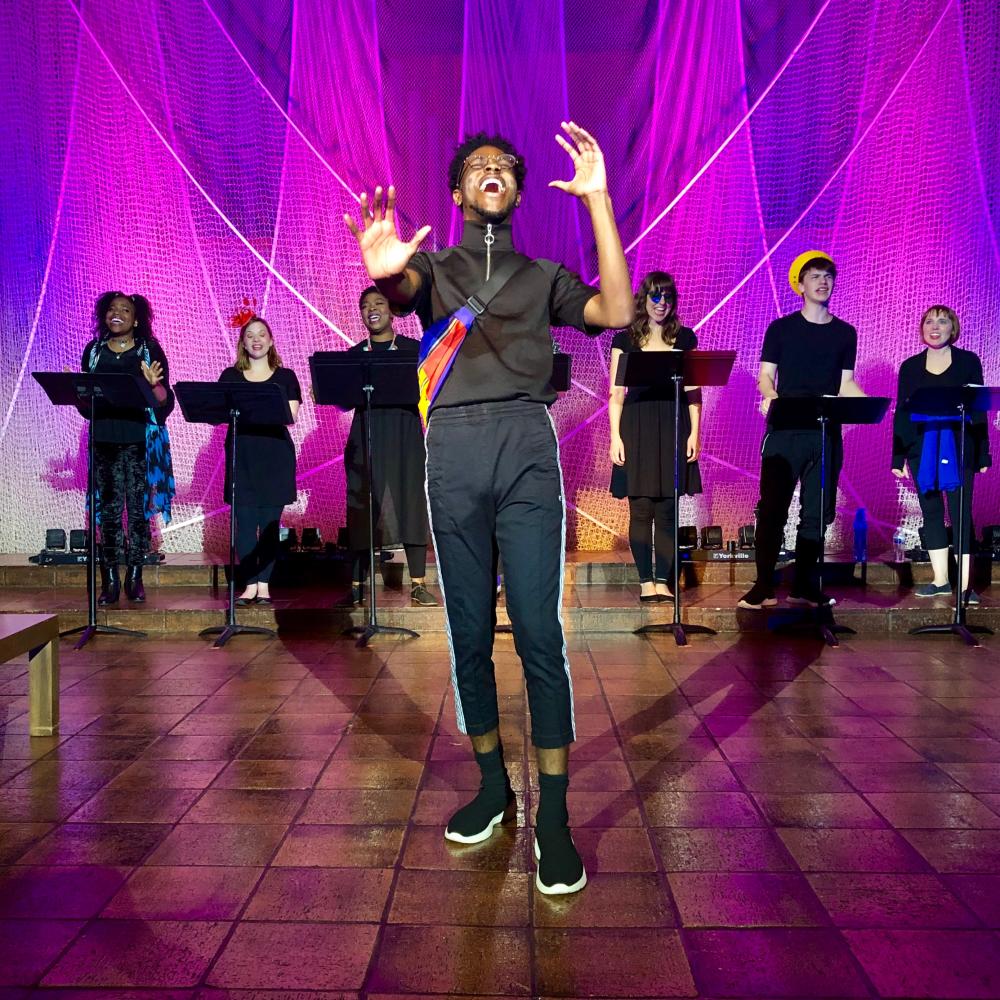 photo of a performance at Trinity Street Playhouse. A man wearing black athletic clothing stands in the center, singing, and a chorus stands behind him. Each chorus member is singing and stands behind a black music stand