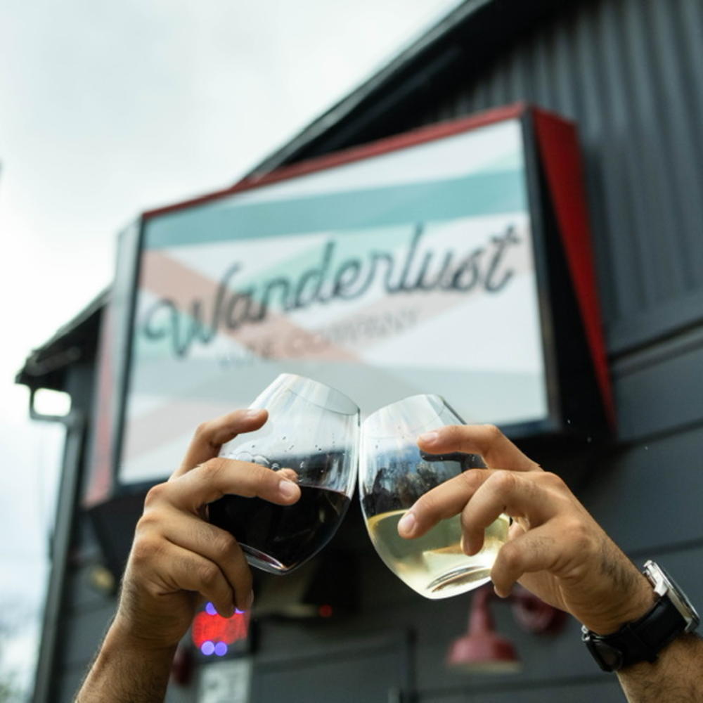 Two hands cheersing red and white wine in front of Wanderlust Wine Co.