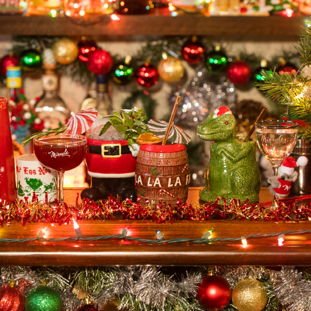 Lineup of quirky holiday cocktail mugs and glasses on decorated bar top.