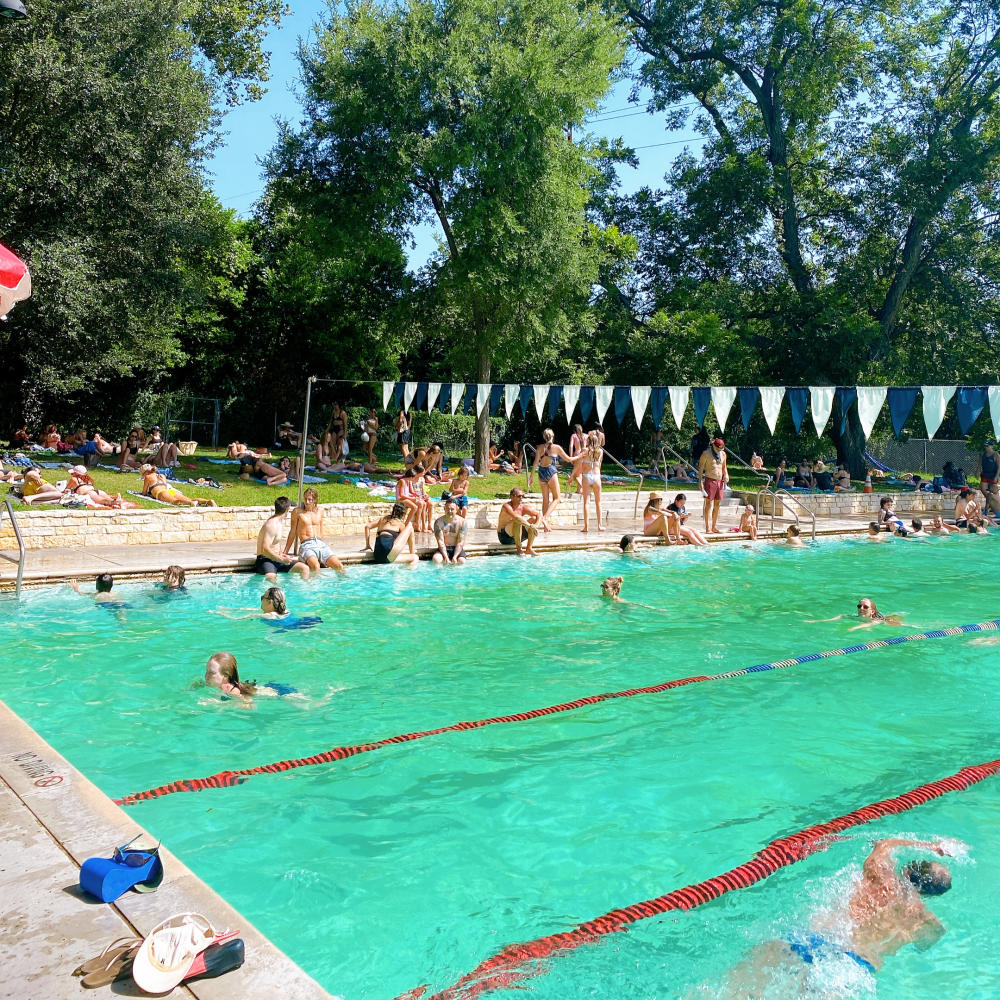 People swimming laps at Deep Eddy Pool, with bystanders sitting along the edge with their feet in the water.