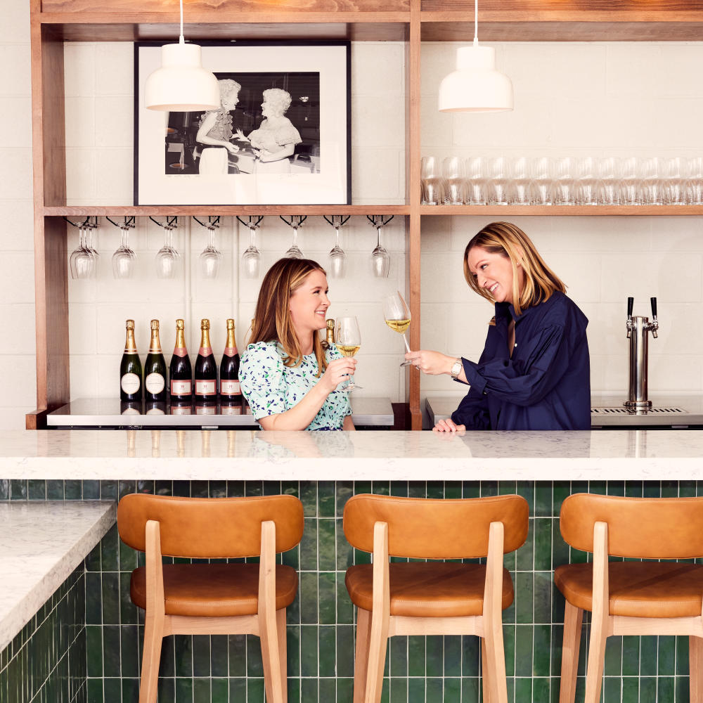 Flo's owners standing behind green-tiled bar with glasses of wine.