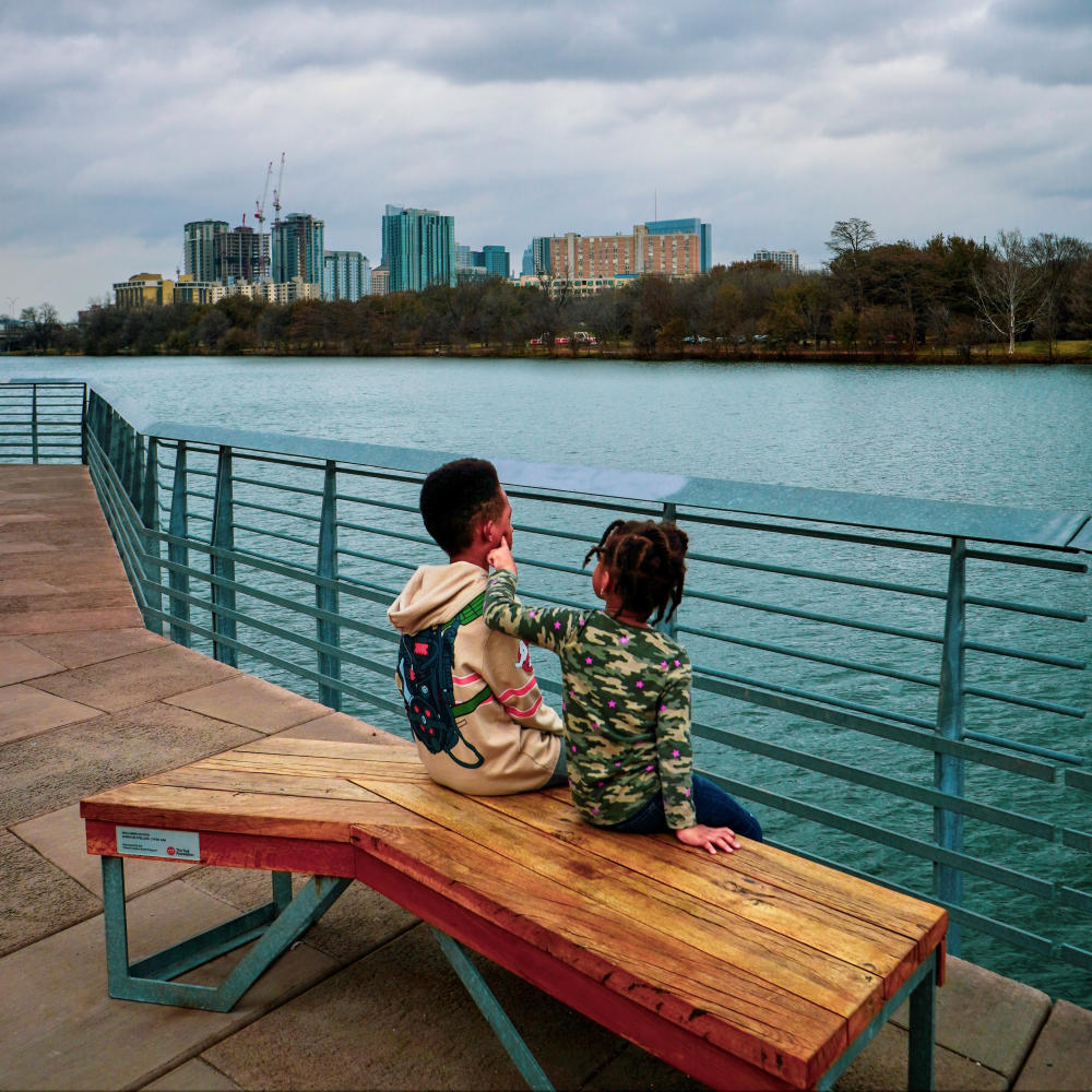 Two young children sitting on a bench on the Hike & Bike Trail Boardwalk. Lady Bird Lake and the Austin skyline are behind them