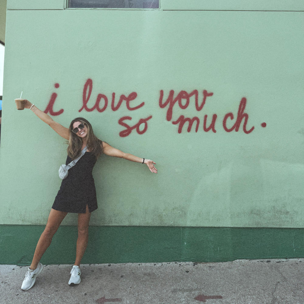 Woman standing with arms wide and iced coffee in hand in front of the iconic "i love you so much." mural.