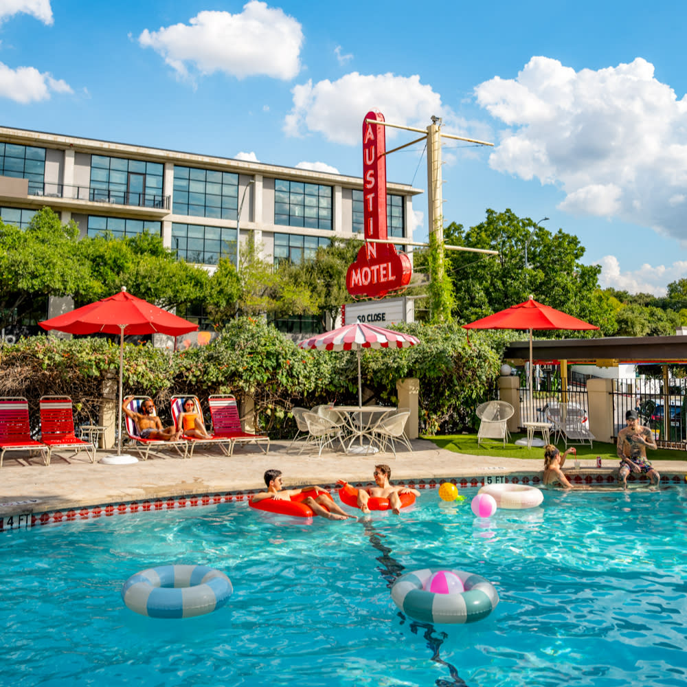 Photo of the pool at Austin Motel during a sunny day, showing the neon sign in the background. Two men sit in inner tubes in the foreground, with another pair sitting on the pool steps behind them, and a man and a woman sitting on the red retro-style lounge chairs under red umbrellas in the back.