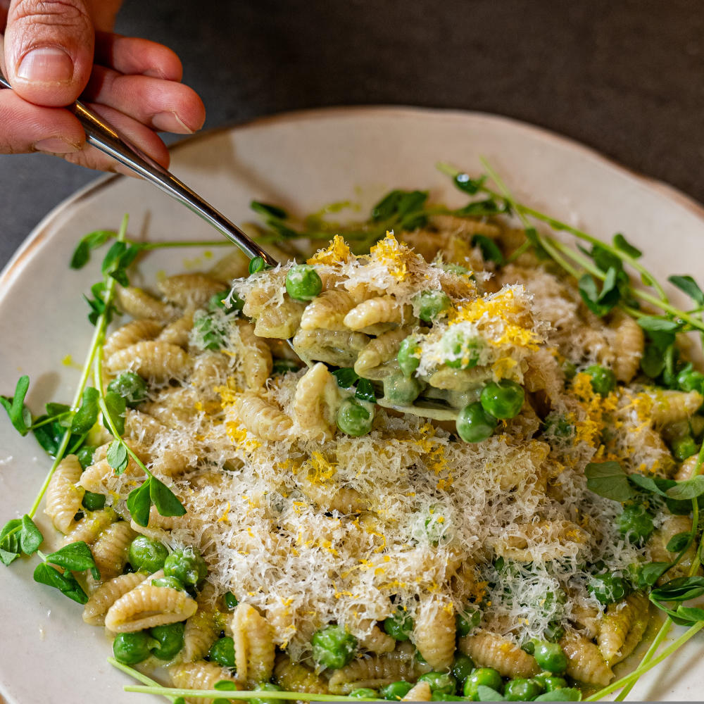 Hand scooping a spoonful of pasta covered in finely shredded cheese, peas and microgreens.