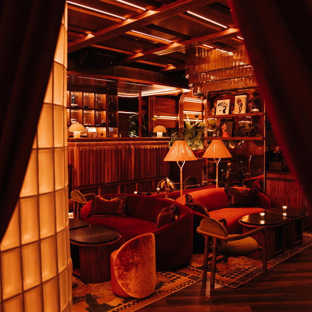 The lounge seating area of Powder Room featuring velvet sofas, lamps, the bar in the background, and deep red color tones filling the whole space.