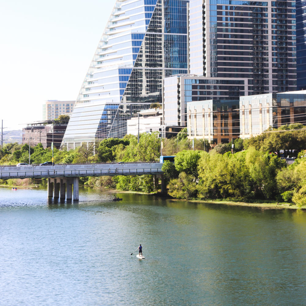 Single paddleboarder on Lady Bird Lake near a bridge and skyline buildings.