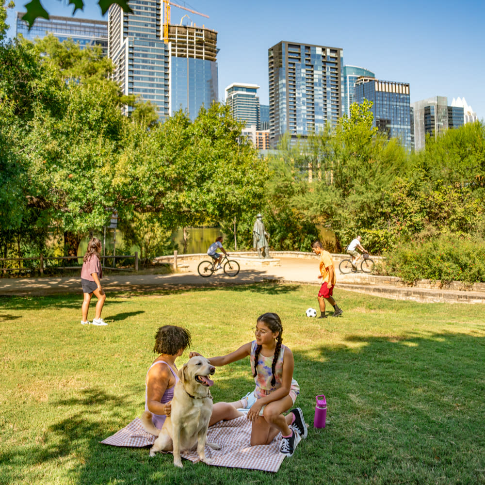 Family on the lawn at Auditorium Shores. A father and daughter playing soccer while a mother and daughter pet a dog on a picnic blanket.
