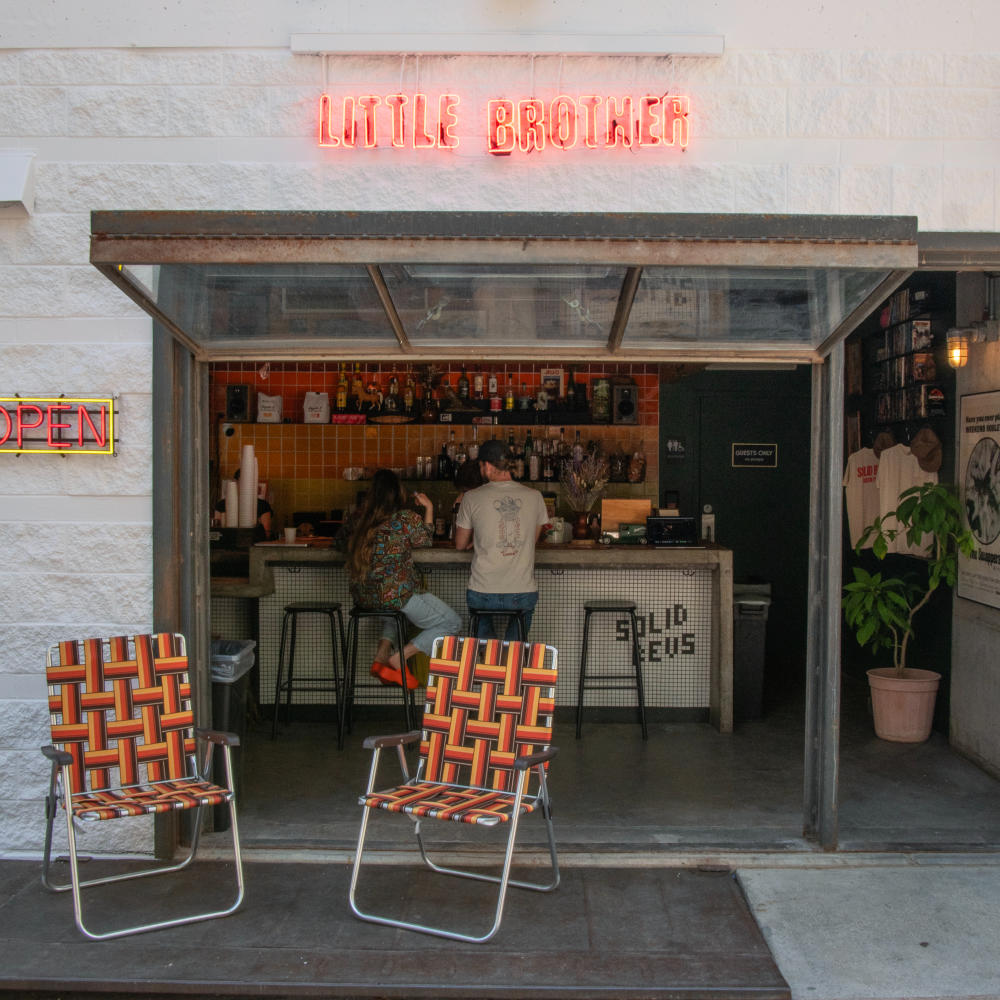 Two people standing inside the open "garage door" of a very small bar with only four barstools. The garage-door style opening has a neon sign "Little Brother" above it.