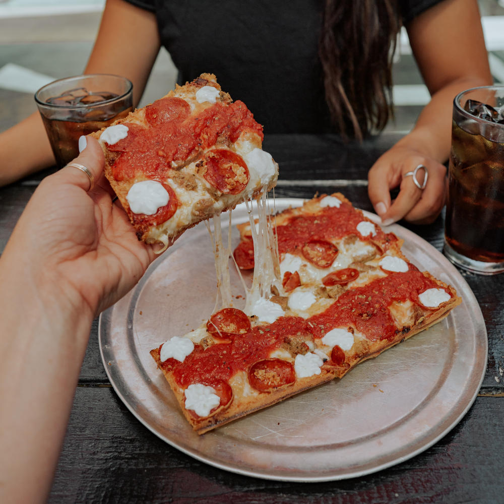 Woman sitting at a table with glasses of coke and a Via313 Detriot style pizza.