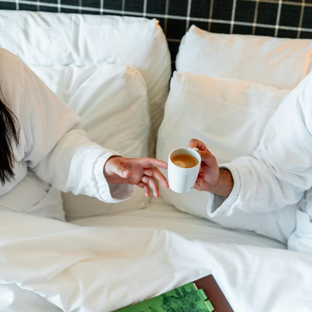 Two people in robes cheersing mugs of coffee in a hotel bed.
