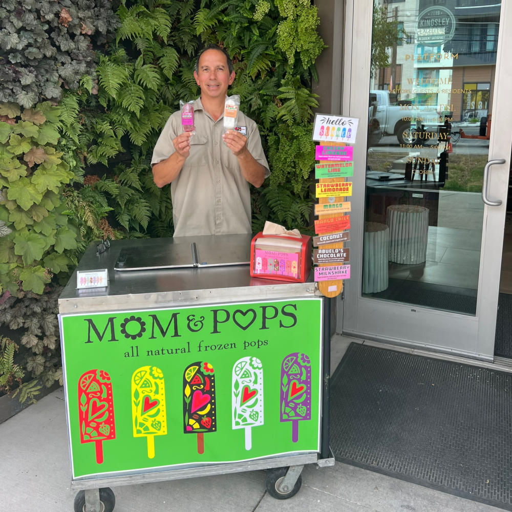 Mom & Pops owner Manuel Flores standing in front of greenery wall with popsicles and paleta cart.