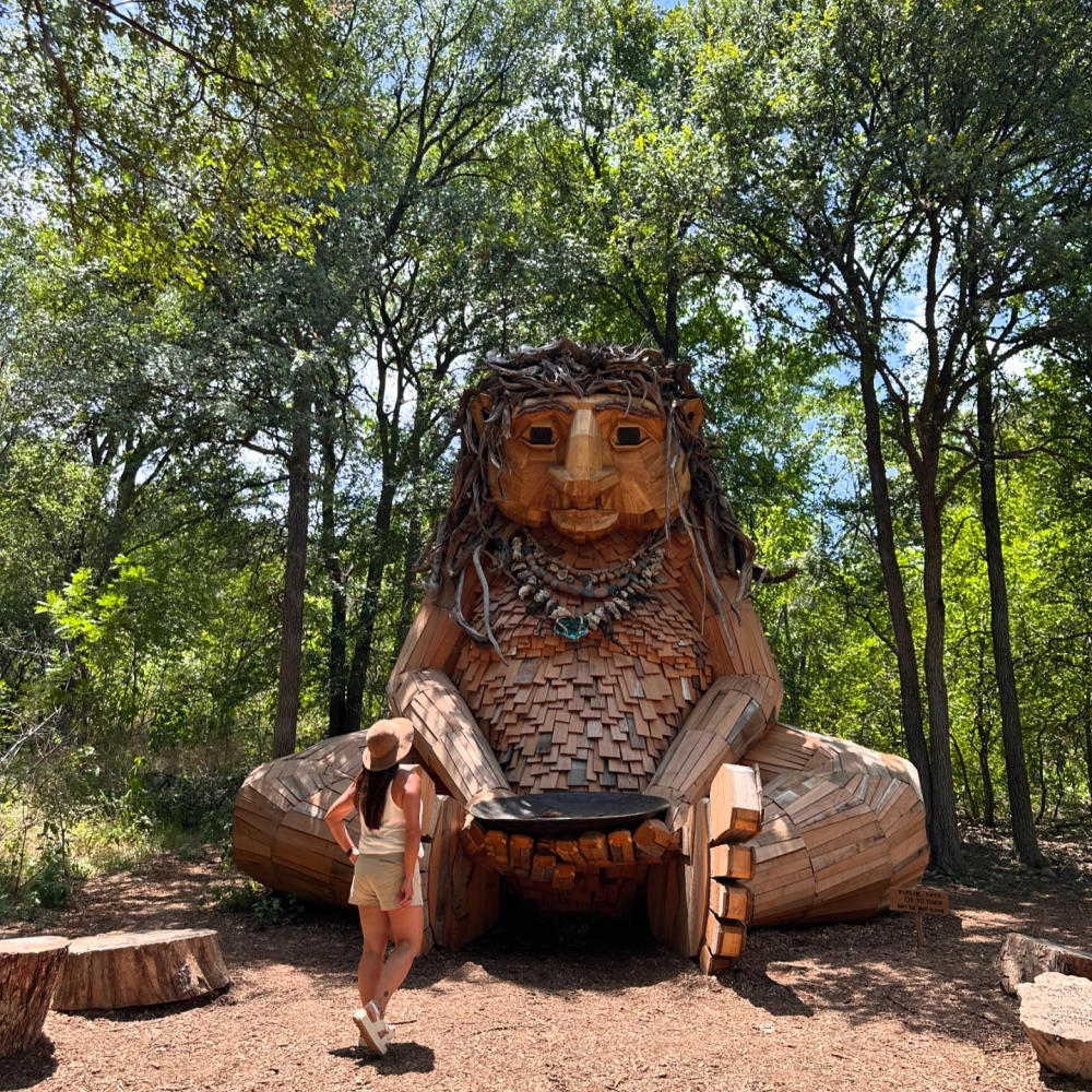 Woman walking up to a giant wooden troll statue up against bright green trees.