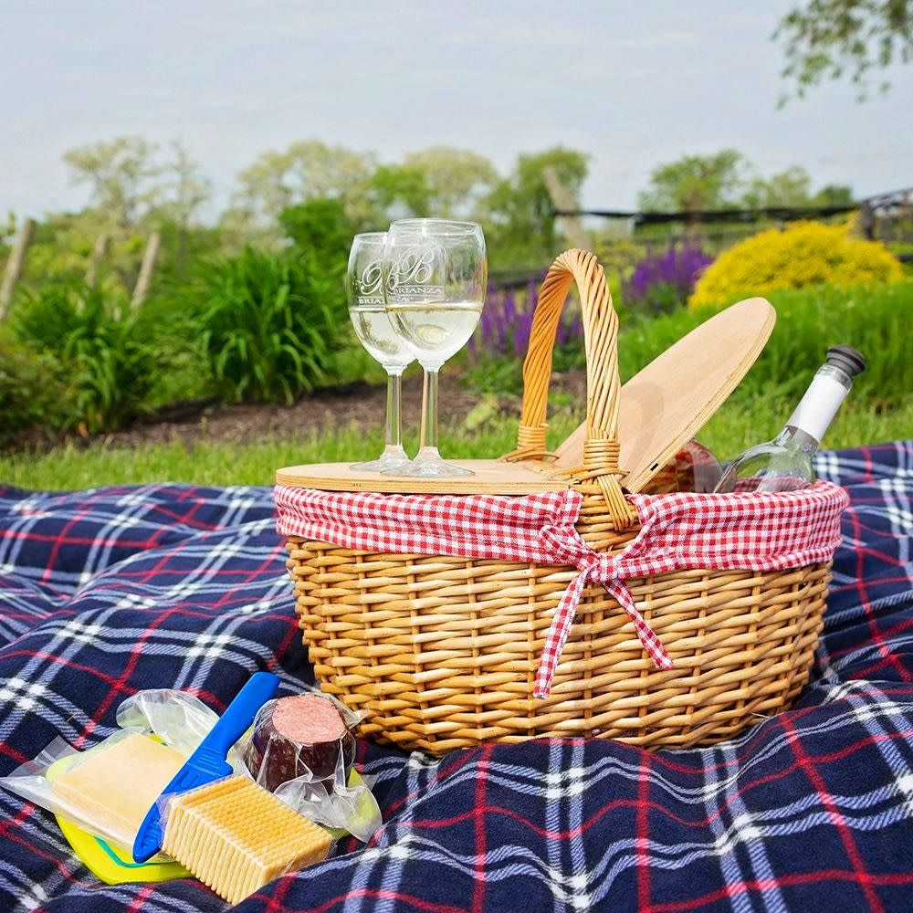 Image is of a picnic basket with a wine bottle coming out of one side, two glasses with white wine on the other and the basket on a plaid blanket outside.