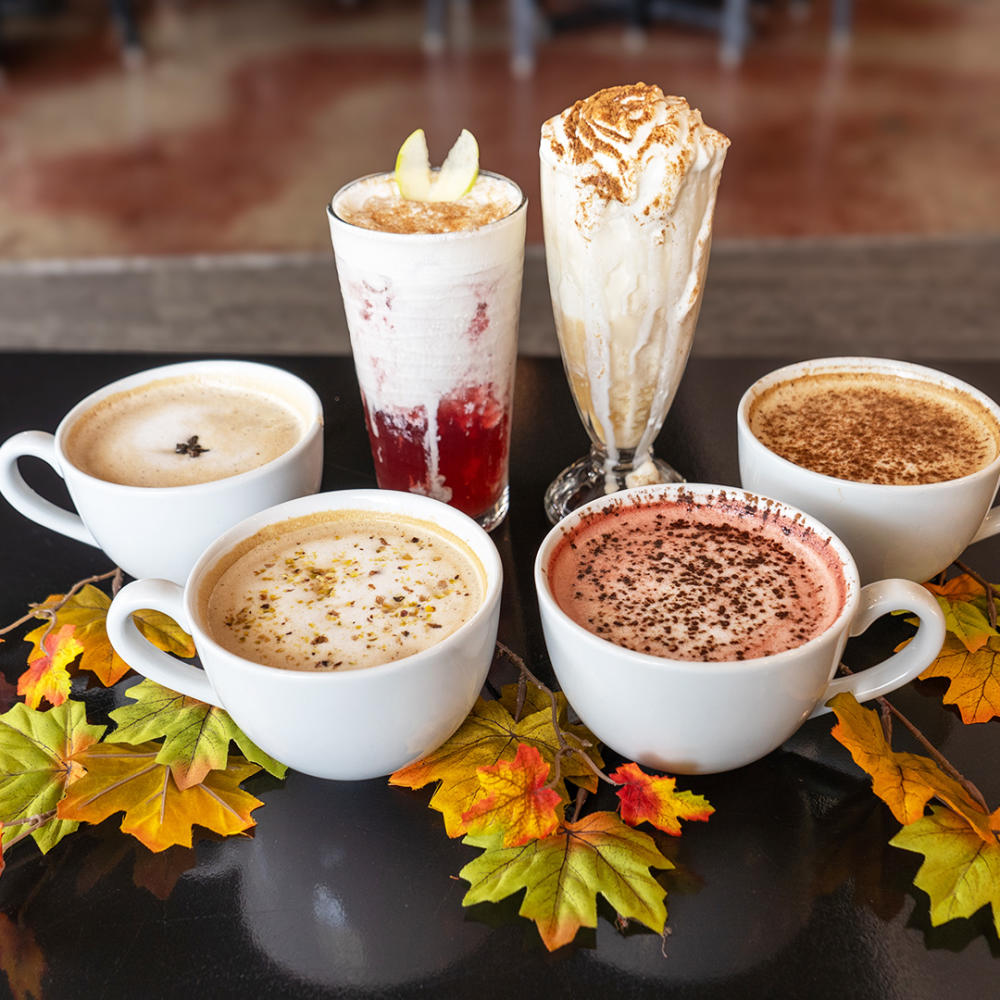 Six fall drinks on a table surrounded by leaves.