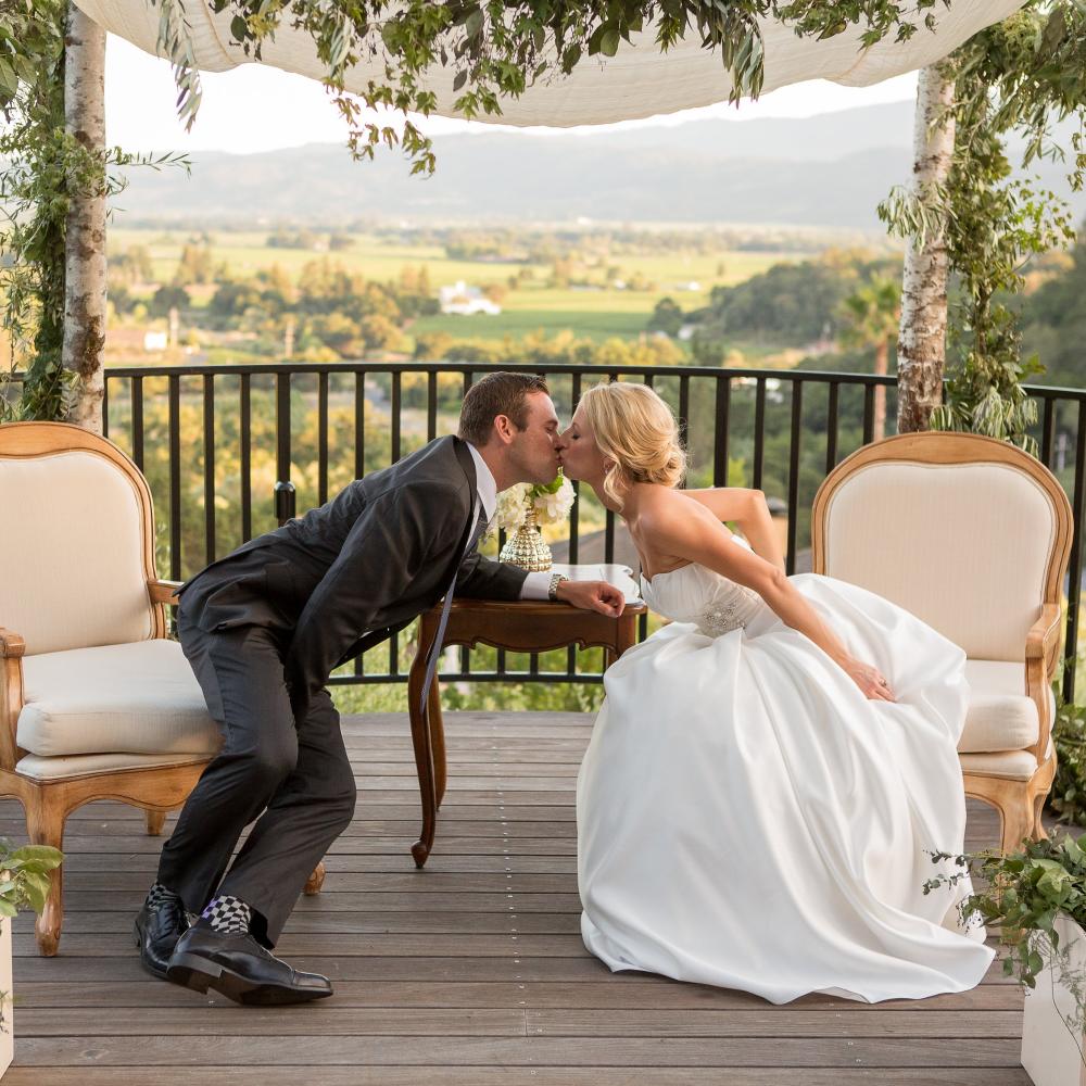 Man and woman sitting in white cloth chairs kissing over a table