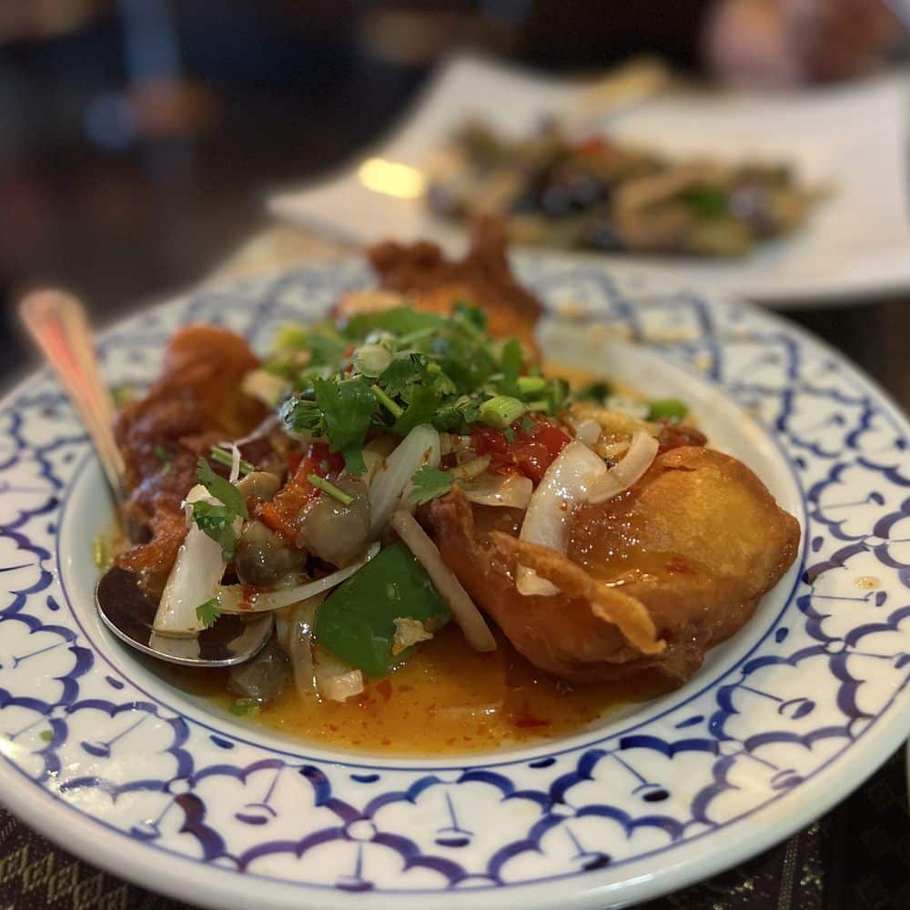A plate of stir-fried tofu with vegetables garnished with chopped scallions, served on a white and blue patterned plate, with a blurred background showing another dish.