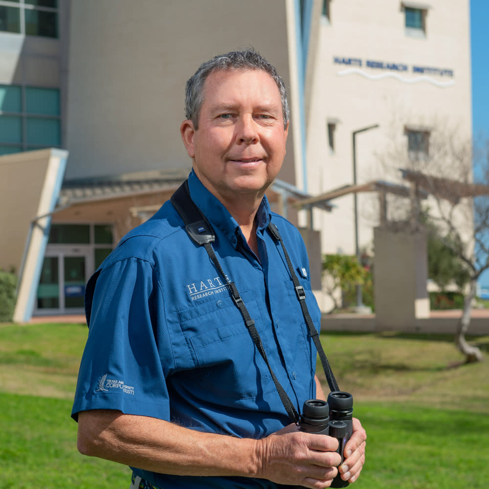A man in a blue button up shirt smiles at the camera. He wears binoculars around his neck.