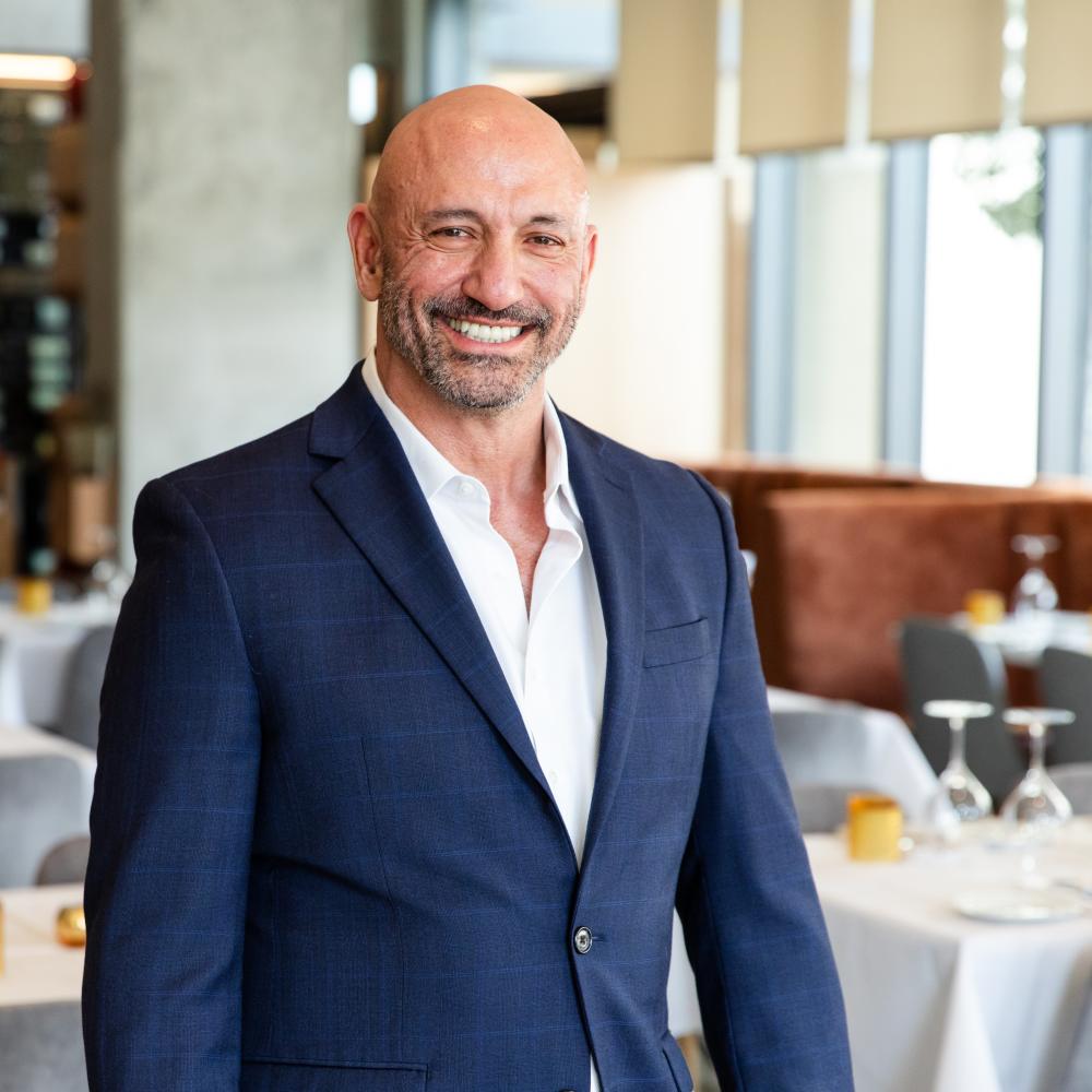Cary Attar (40-50s) is wearing a navy suit with a white button-down shirt. He is fit, bald, and has a short beard. He is standing in Fielding's Local Kitchen + Bar, where the tables behind him are set with white cloths, yellow candles, and upside-down wine glasses.