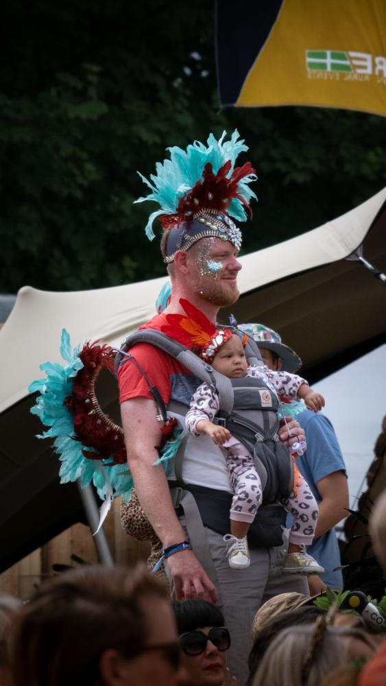 A man with glittery face paint and feathered headdress carries a baby in a leopard onesie at a festival. Both wear matching accessories, atmosphere is festive.