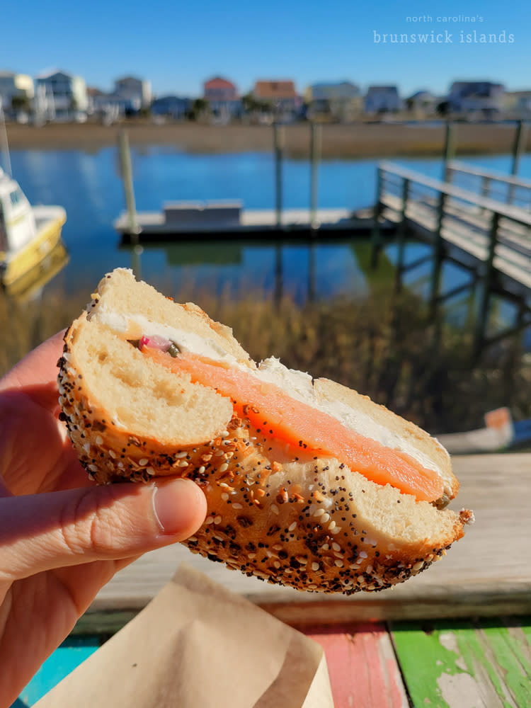 A hand holding an everything bagel with lox and cream cheese with a view of a canal in the background
