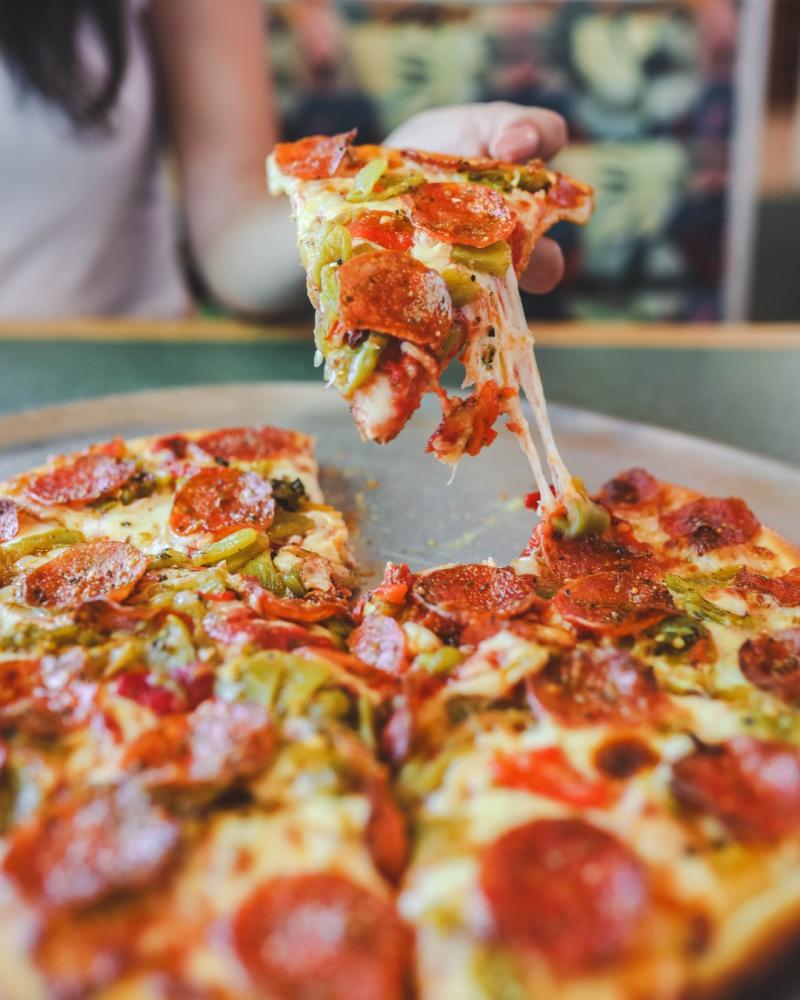 Someone pulls a slice of pizza away from the main pie. The pizza is pepperoni and green chile.