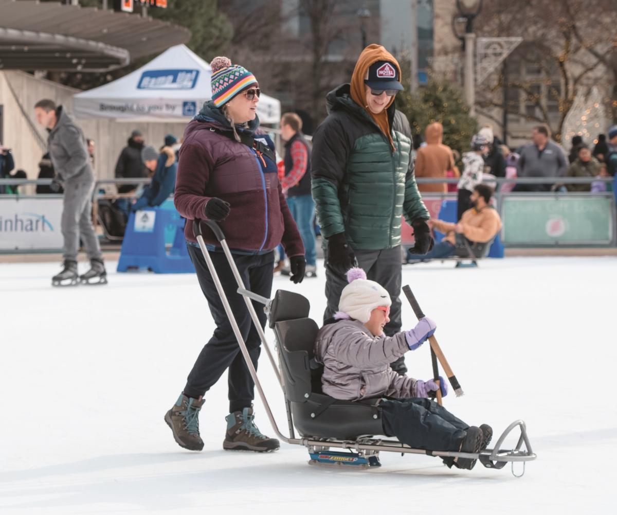 Two adults accompany a child using an adaptive ice-skating sled at the Slice of Ice rink in Red Arrow Park.