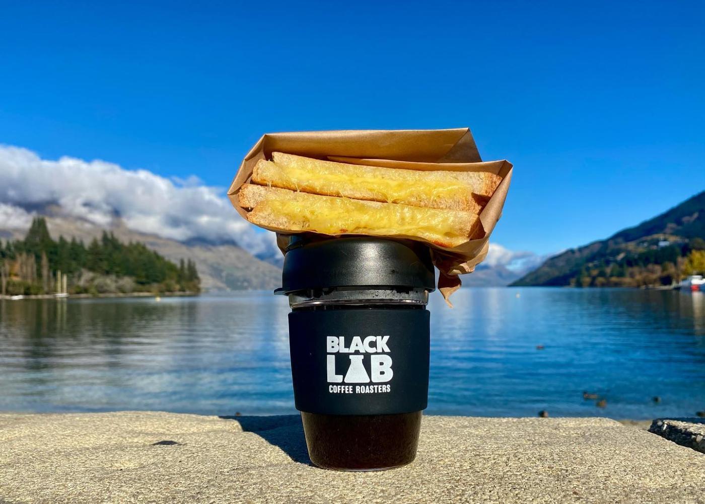 Resuseable coffee cup with whit weiting that reads Black Lab Coffee Roasters with cheese toasty sitting on top of the cup and a view of lake and mountains in the background