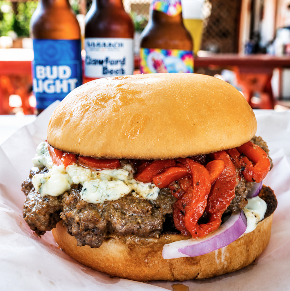 A Fourth of July Burger at Lankford's features a thick beef patty, roasted red peppers, red onion slices, ranch dressing, and blue cheese crumbles on a brioche bun. Bud Light Beer bottles stand in the background.