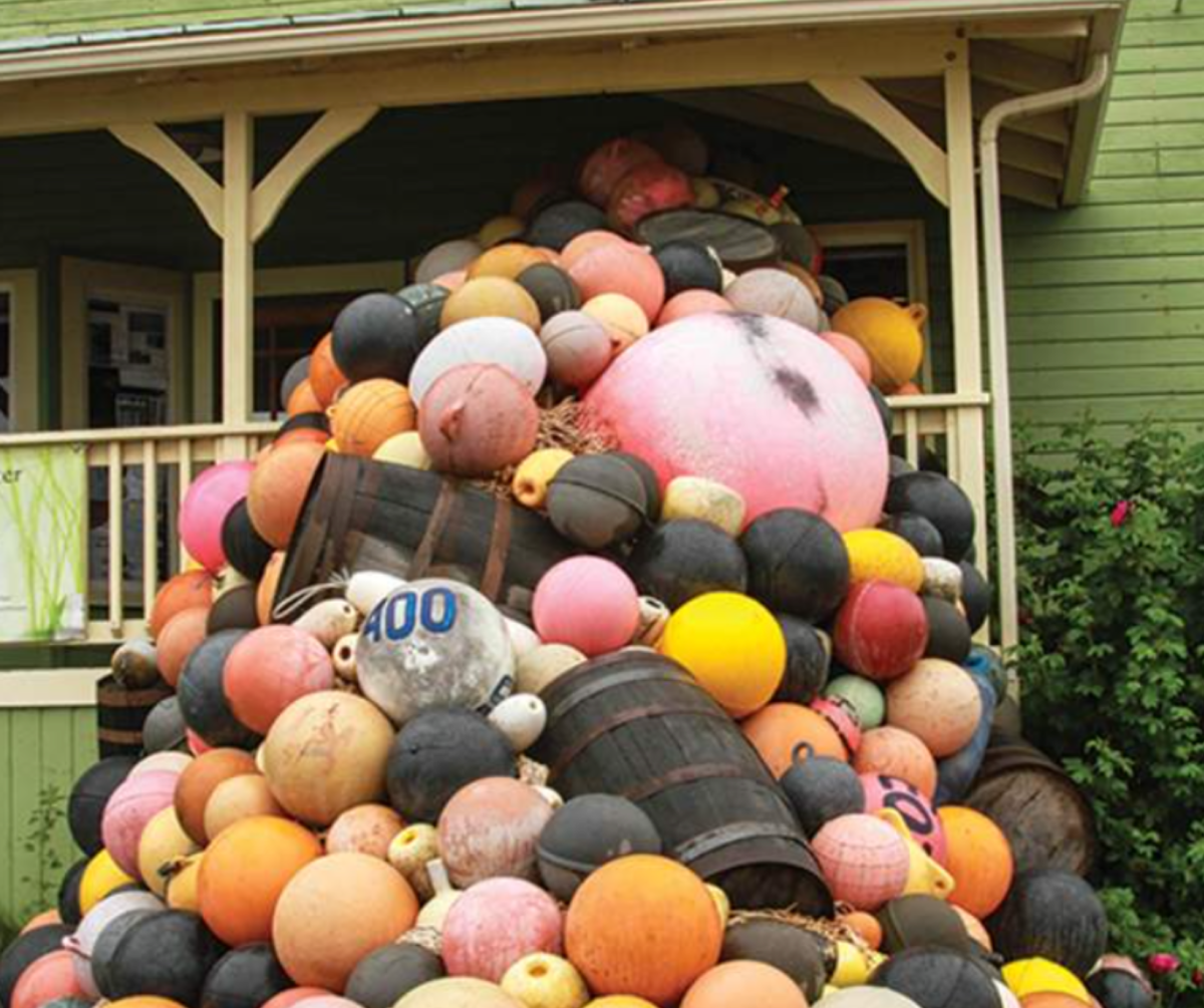 A large, colorful stack of fishing buoys and barrels are piled high in front of a charming green house, creating a vibrant and playful display of maritime life in Homer.