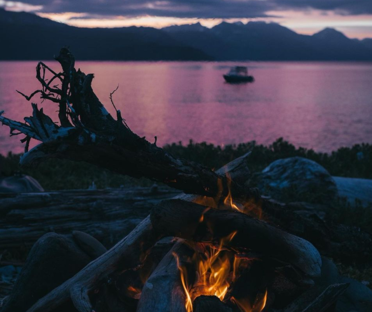 A cozy beach campfire burns warmly in the foreground, with a serene, purple-hued sunset over the mountains and water in the background, capturing a peaceful Alaskan evening.