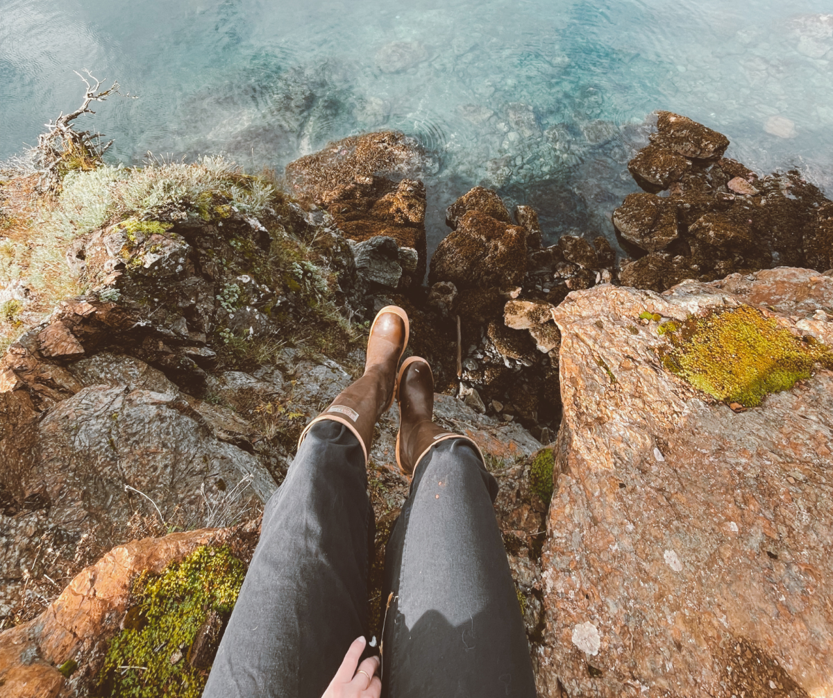 A person’s legs, dressed in dark pants and wearing brown boots, dangle over a rocky ledge overlooking crystal-clear blue water below, showcasing a tranquil moment by the shore.