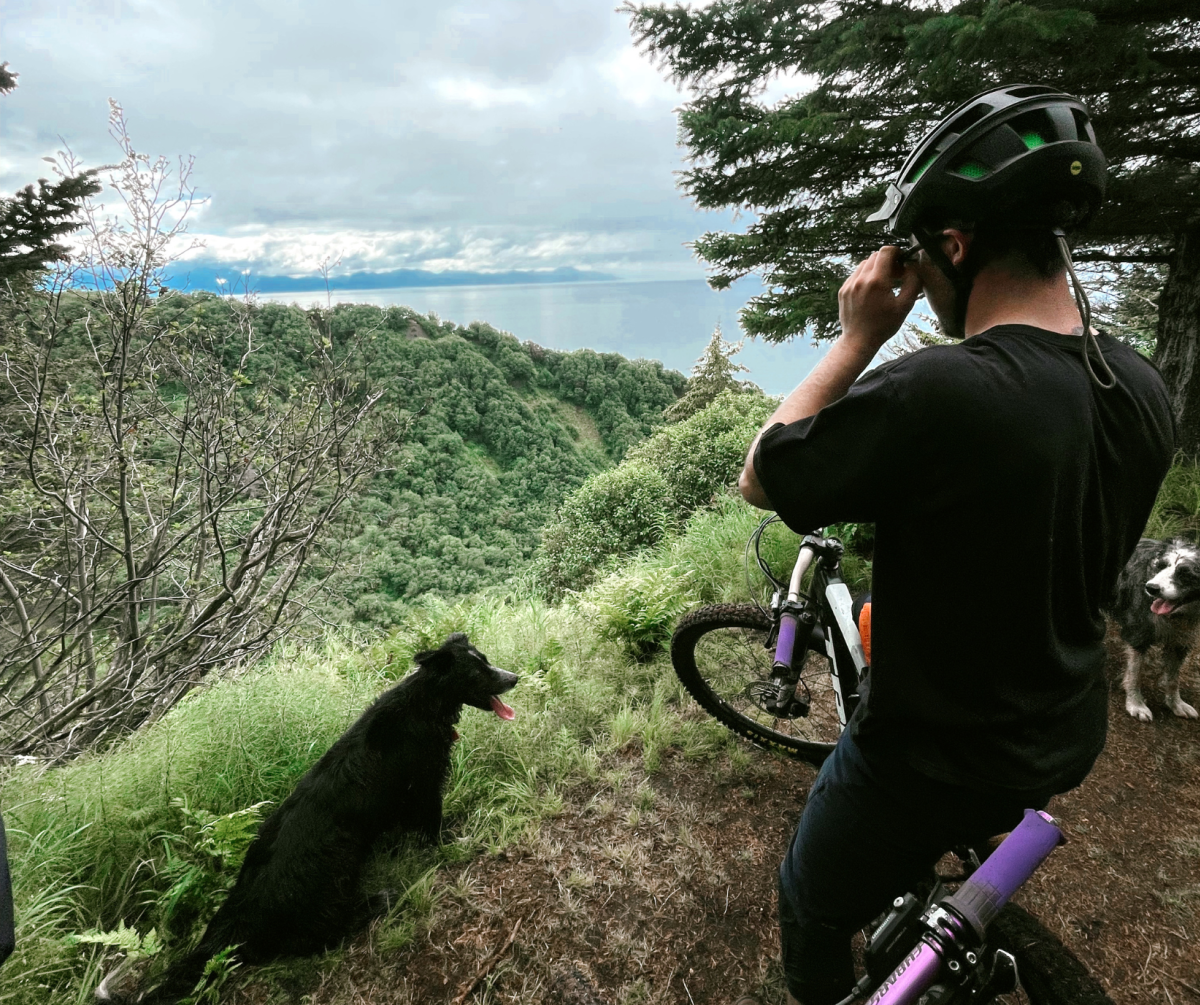 A person in a helmet and black t-shirt prepares for a mountain biking adventure on a trail overlooking a lush green valley with expansive views of the water and distant mountains. Two dogs stand nearby, ready to join the adventure.