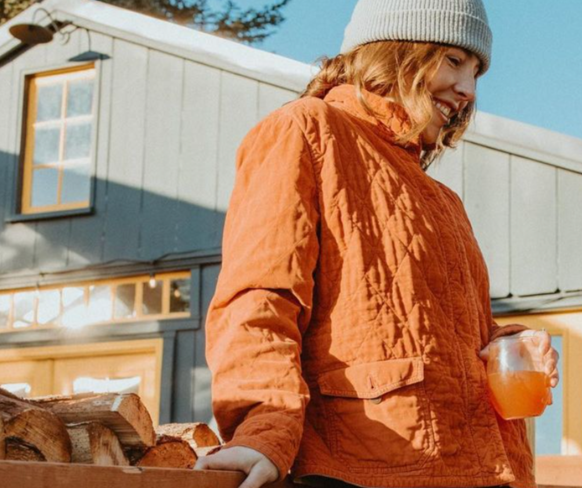 A woman in an orange jacket and gray beanie carries a load of firewood with a smile on her face, holding a beverage in her other hand, enjoying a crisp day in Homer.