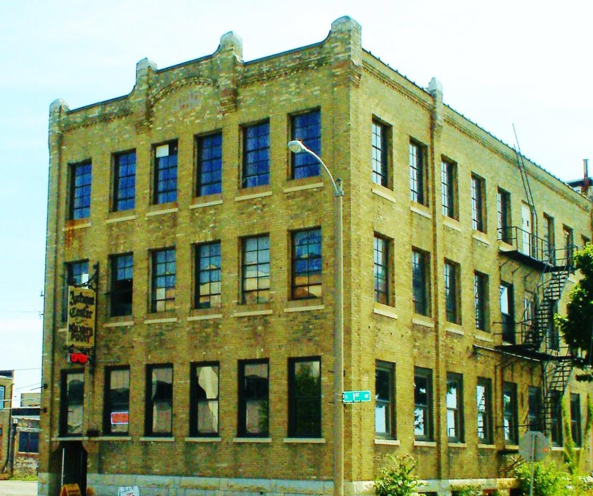 The Antique Center of Walker's Point occupies a historic three-story cream brick building on a street corner. Tall industrial windows, a painted hanging sign and an exterior fire escape highlight the building's warehouse architecture.