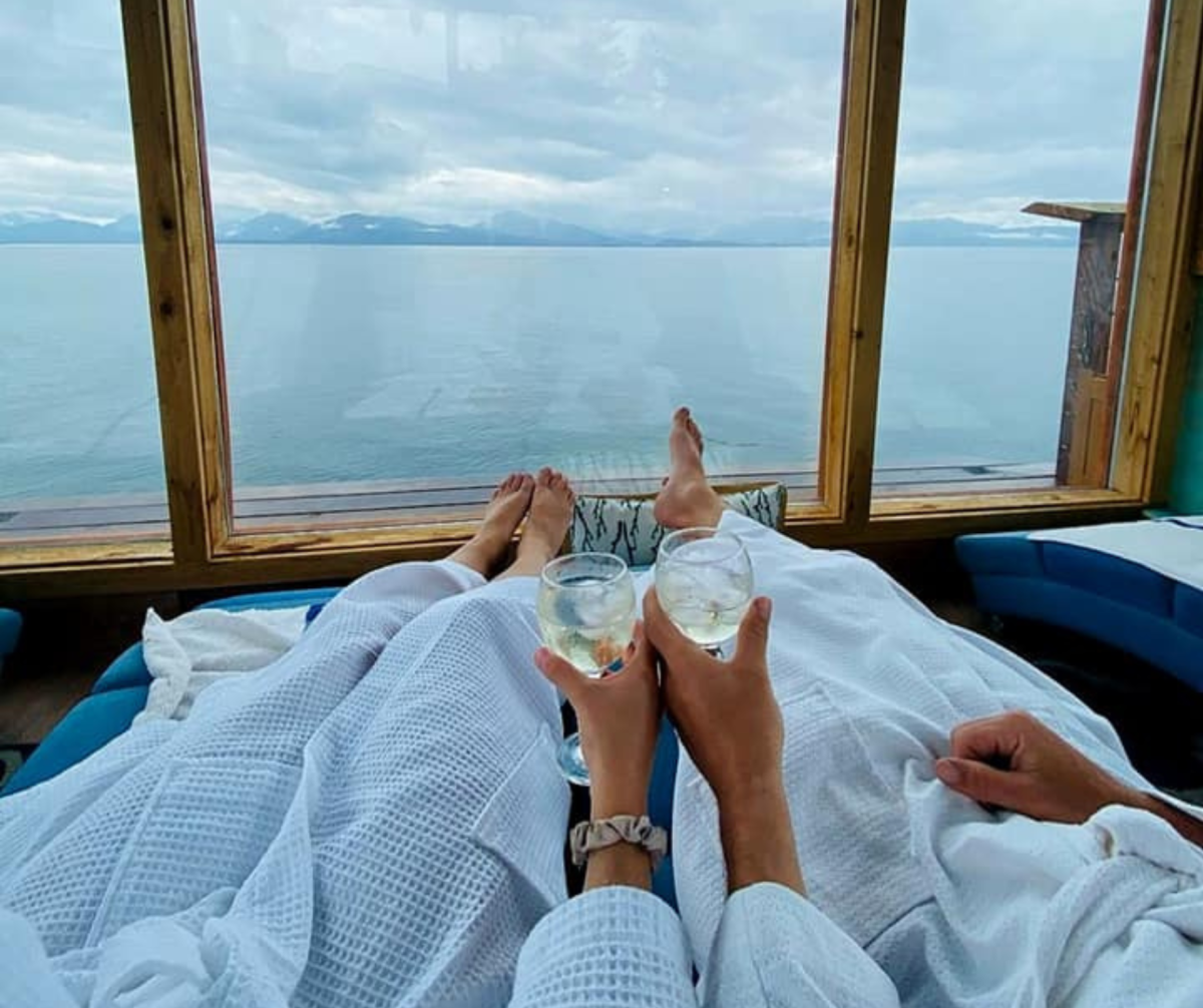 Two people relax in plush white robes while enjoying glasses of white wine, overlooking a breathtaking view of calm ocean waters and distant mountains through large windows in a cozy room. The serene setting creates a peaceful and luxurious retreat vibe.