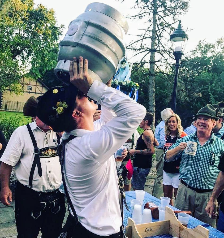 A man in traditional German clothing tipping up a beer keg to his mouth while a smiling crowd looks on at Mainstrasse Village Oktoberfest