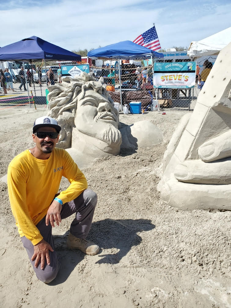 A man in a yellow long sleeve shirt kneels down next to a sand sculpture depicting a man's face and hand coming up out of the ground, rocking out on a guitar