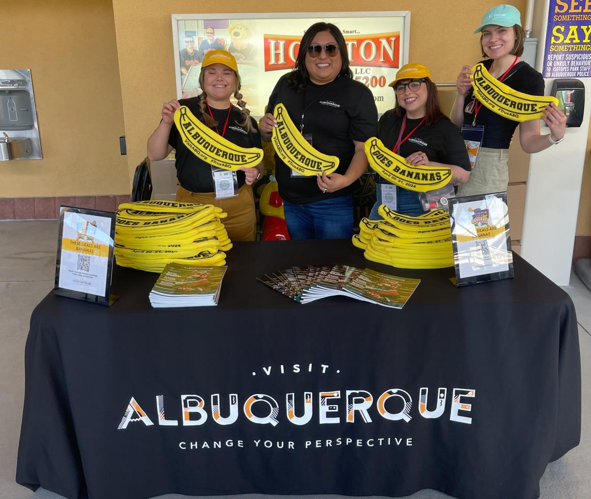 Four Visit Albuquerque employees hold up Savannah Banana hats at the Visit Albuquerque table.