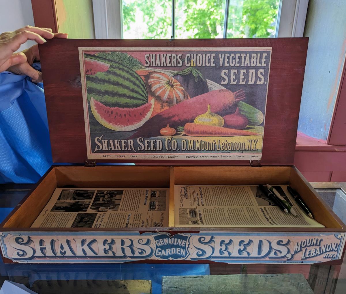 Image is of a Shaker Seed box that is made of wood and say's "Shaker Seeds" with magazine articles inside.
