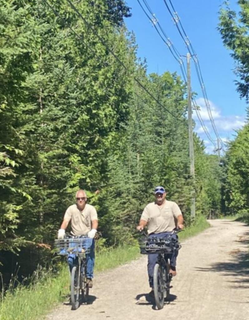 Two electrical line foremen ride bicycles with tool carts on a dirt path beneath overhead power lines on Mackinac Island