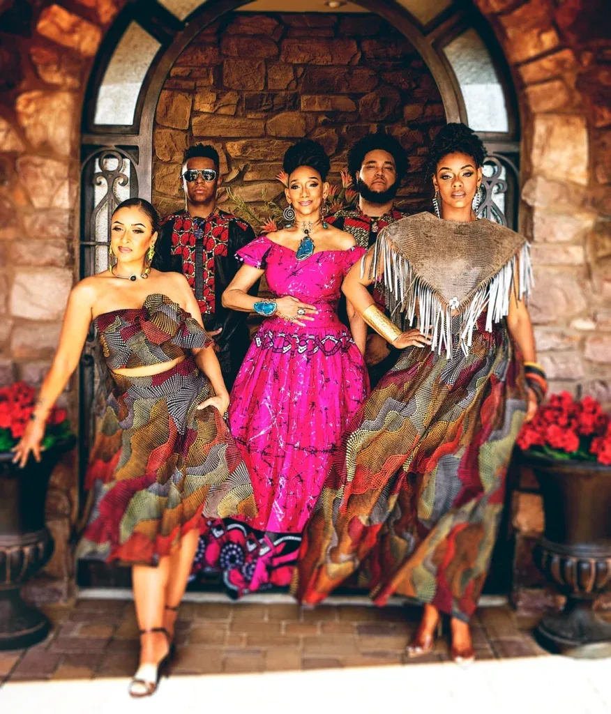 Five people dressed in colorful traditional outfits posing confidently in front of a rustic stone archway, with warm tones and vibrant patterns highlighting their garments.