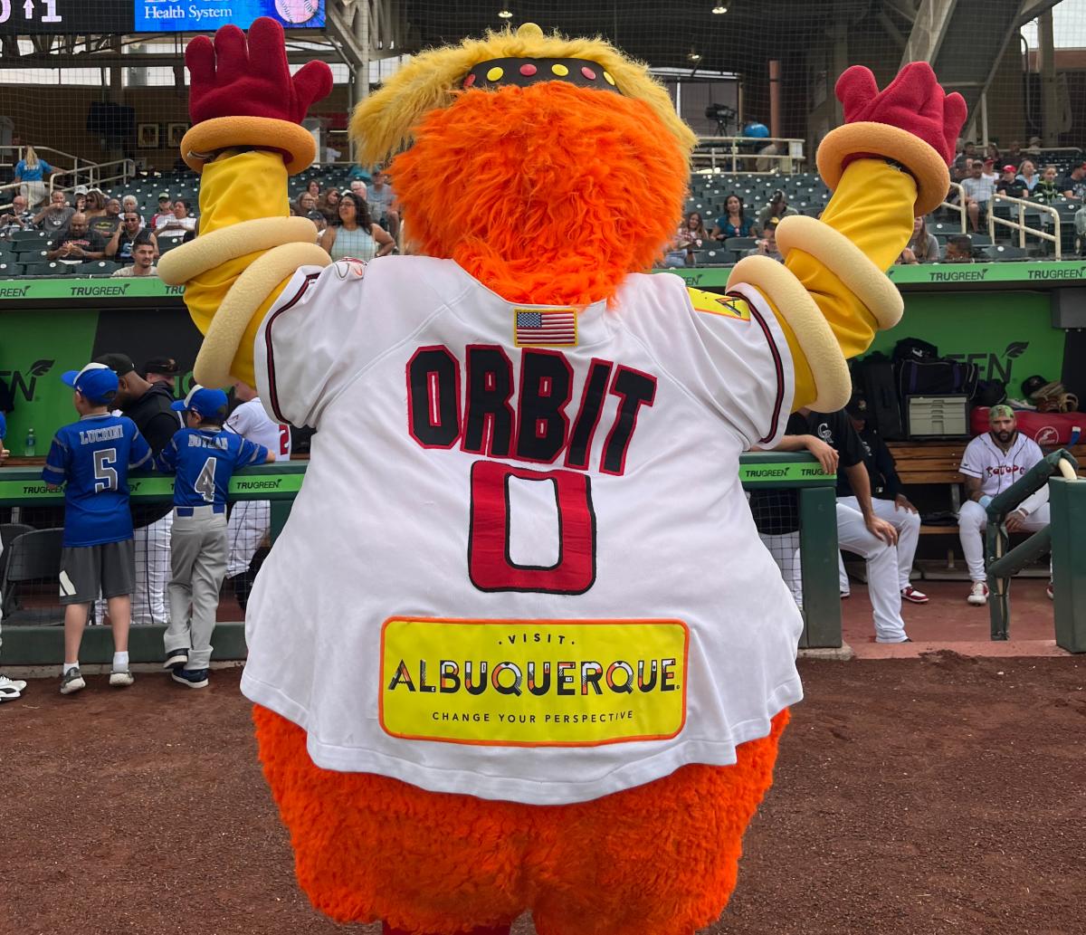 Orbit holds up his hands to the stands at Isotopes Park. His jersey includes a bright yellow Visit Albuquerque patch.