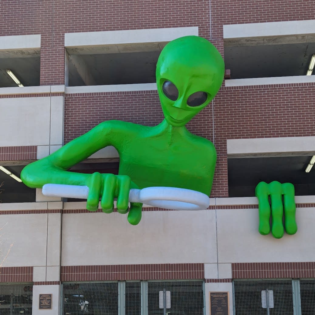 A sculpture of a 30 foot green alien leaning out of a parking garage and peering through a magnifying glass