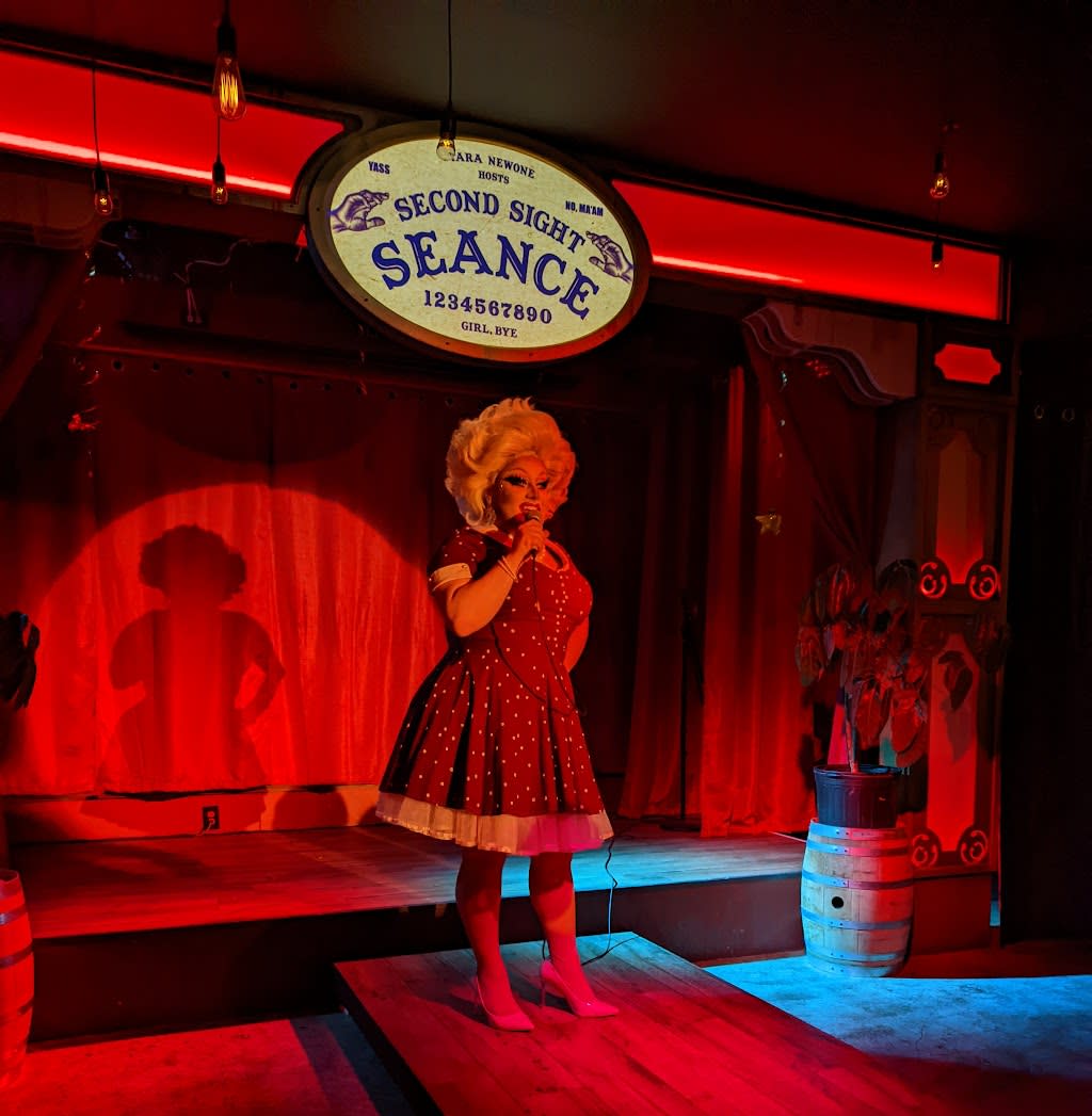 Drag Queen on the stage of Second Sight Spirits with a sign that says Second Sight Seance